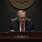 Fed economist at a podium with the Fed seal, in front of screens and tickers during the FOMC.