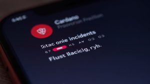 Photorealistic close-up of a Cardano wallet app screen with a red paused badge, shield icon, and fading seed phrase fragments.